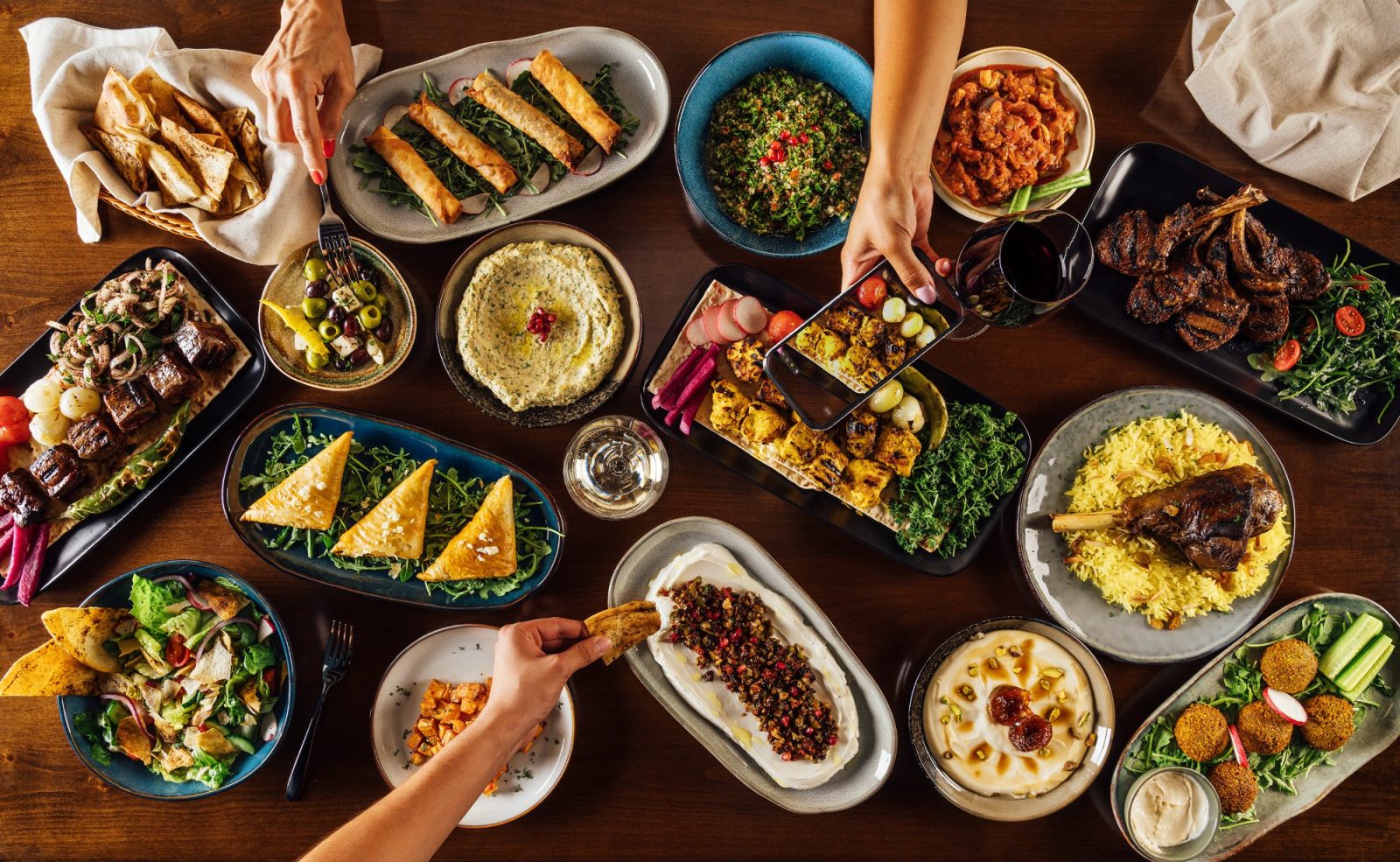 Overhead food photography of a Mediterranean feast at Zeitouna restaurant in Mesa, Arizona, with hands reaching for mezze dishes including hummus, tabbouleh, falafel, kabobs, and lamb shank — photographed by Timothy Fox, Phoenix food photographer