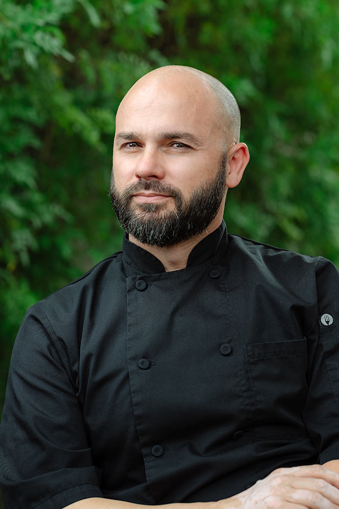 Headshot of a chef wearing a black apron standing in front of green shrubs