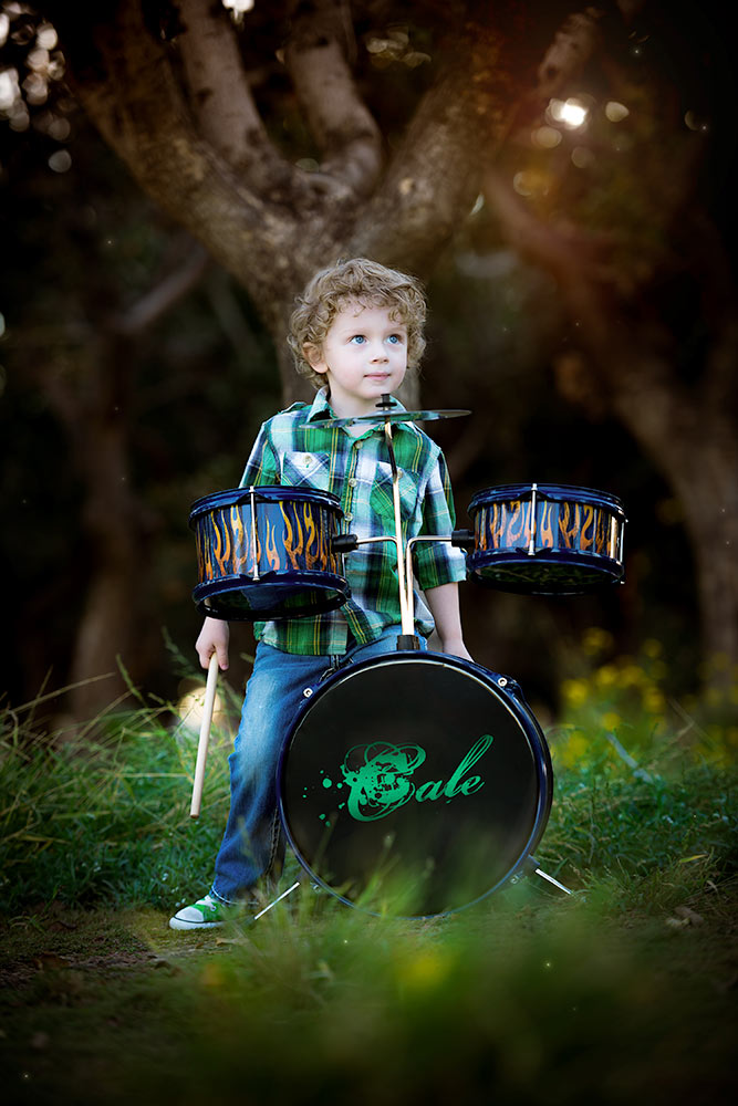 A little boy behind a set of drums with his name on the kick drum in a forest setting
