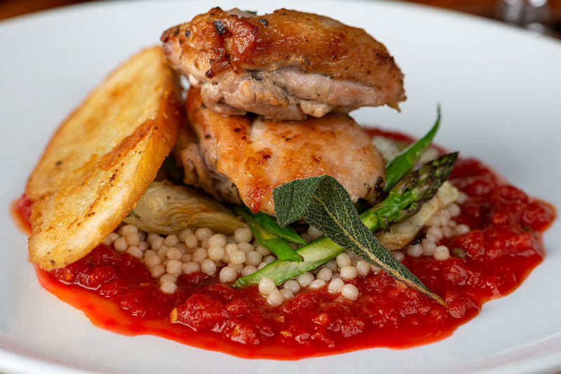 Chicken sitting on a bed of couscous draped in greenery on top of a red sauce