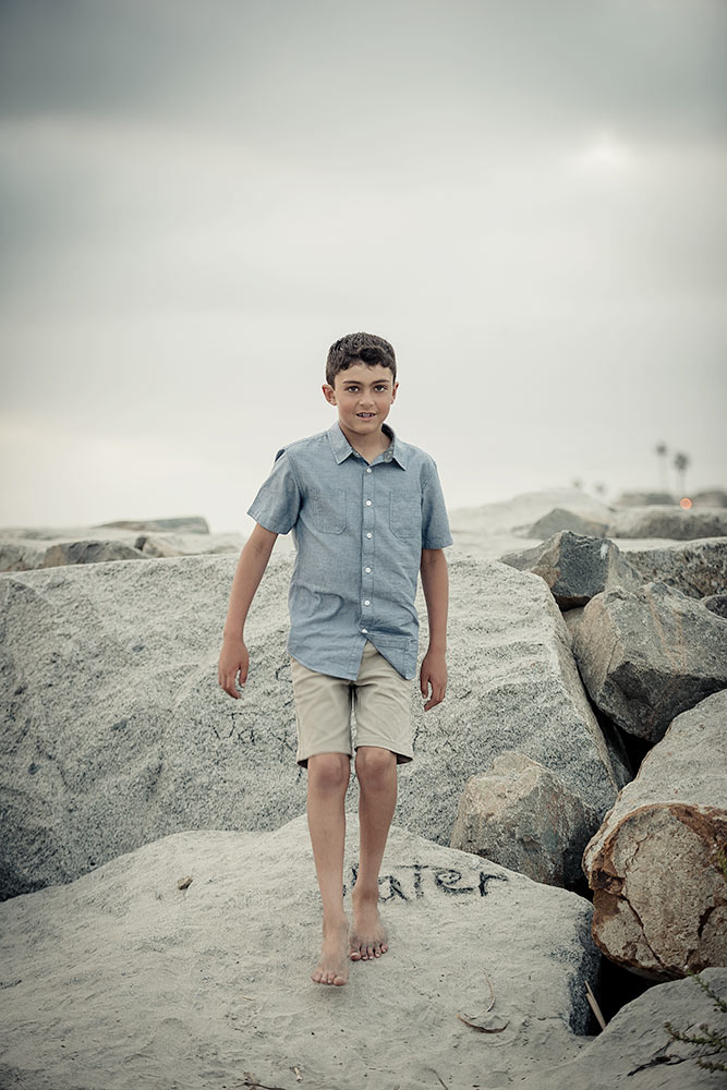 Young boy wearing blue short sleeved shirt and khaki pants walking along the rocks on the beach