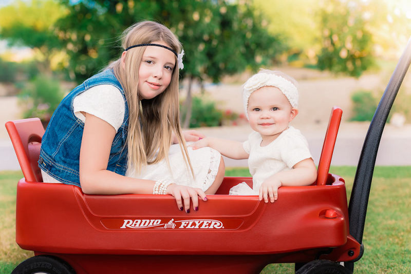 2 little girls pose for a photo in a red wagon in the park