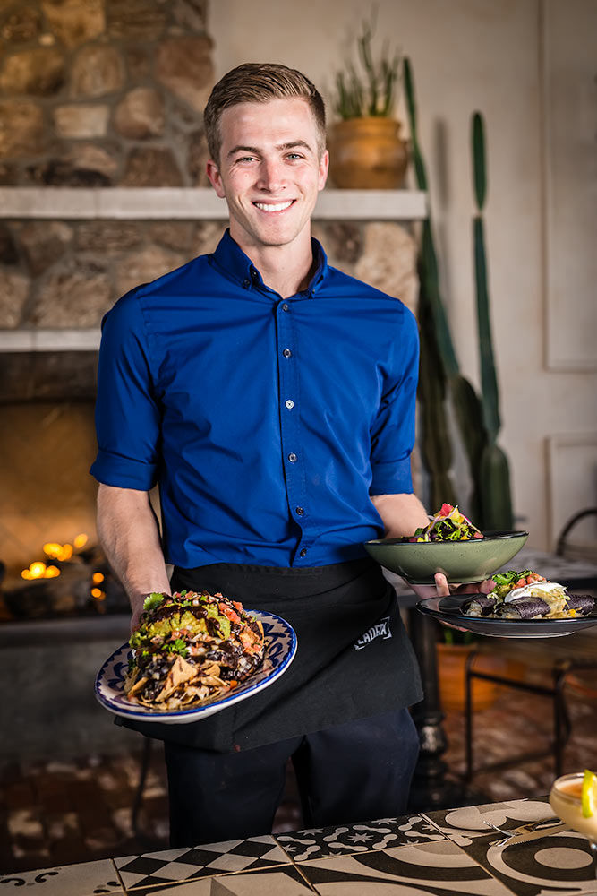 Smiling server with a blue shirt and black apron holding Mexican food plates in front of a fireplace