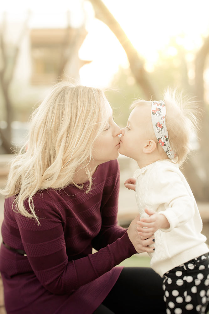 Blonde mother in a fuchsia outfit kneeling to kiss her baby daughter who is wearing a white shirt and pokadot pants