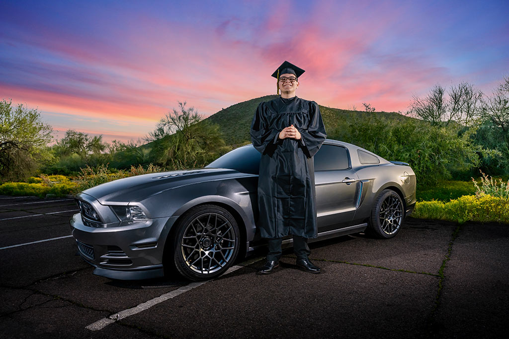 Senior picture with stylish attire, posing in front of a stunning car amidst the desert backdrop, accentuated by a dramatic sunset