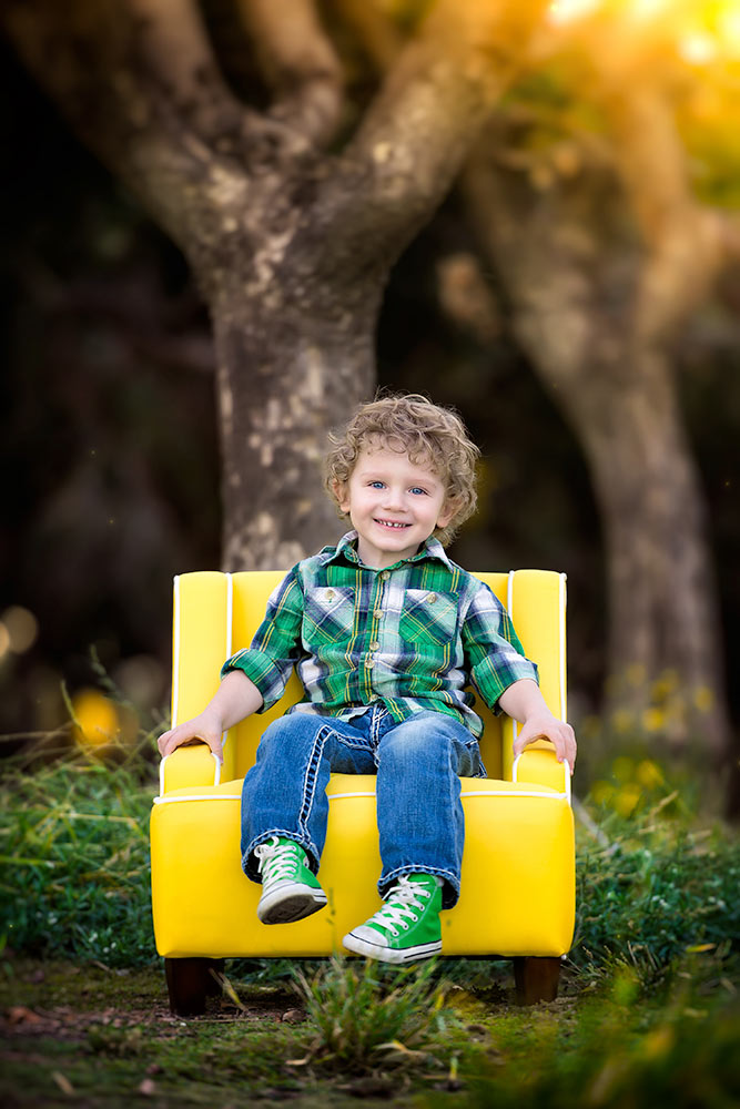 Young boy dressed in a plaid shirt and blue jeans with green shoes sitting on a yellow chair in the forest during golden hour