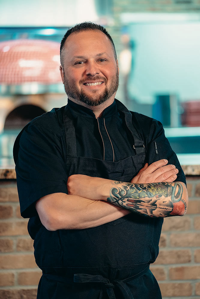 Chef of a restaurant posing with arms crossed with tattoos and a wood fired grill in the back